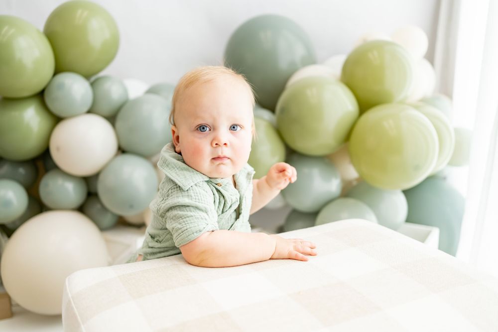 Sweet baby in light green shirt poses against pastel balloon arch backdrop for milestone photo session.