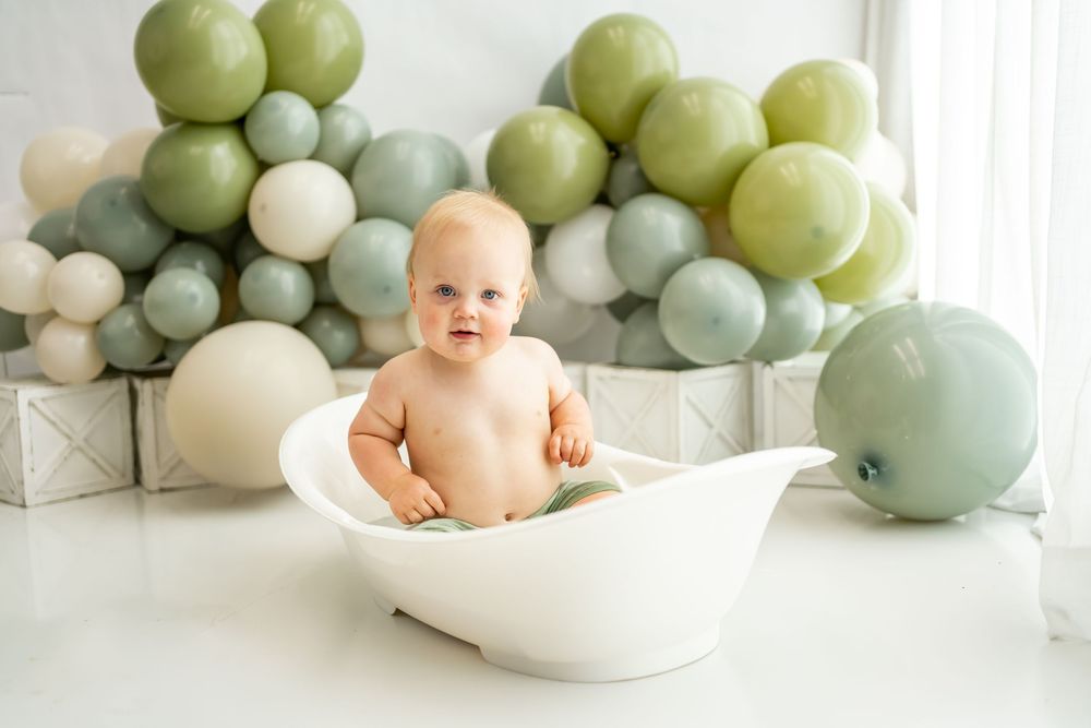A baby sits in a vintage style white bathtub with green and white balloon decorations in the background.