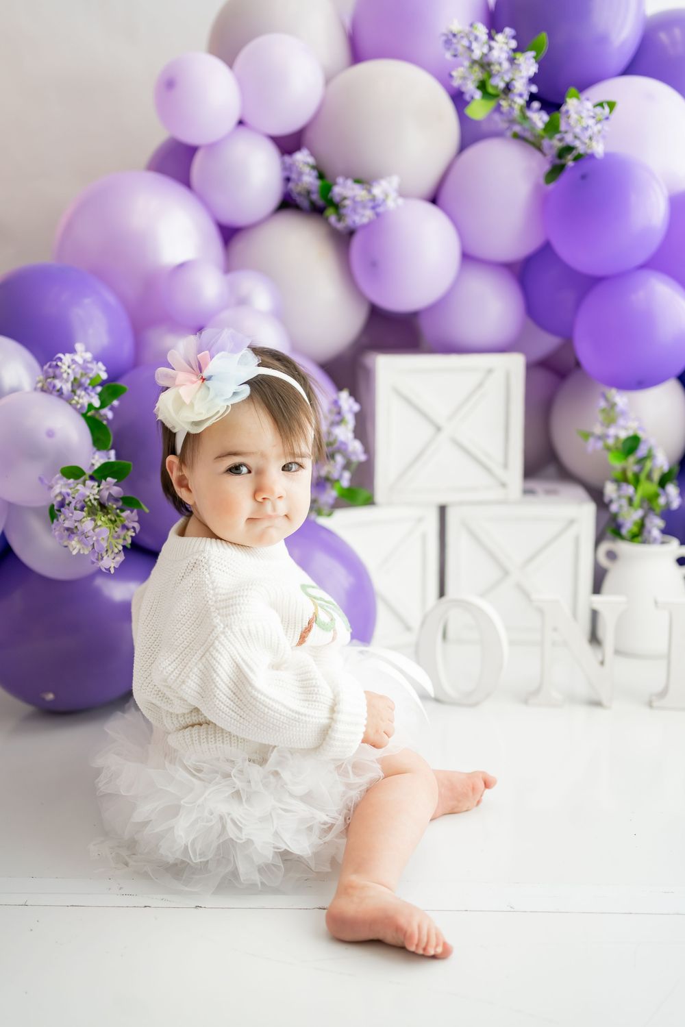 Elegant white and purple balloon arch with lilac flowers decorates a dreamy studio setup with decorative white boxes.