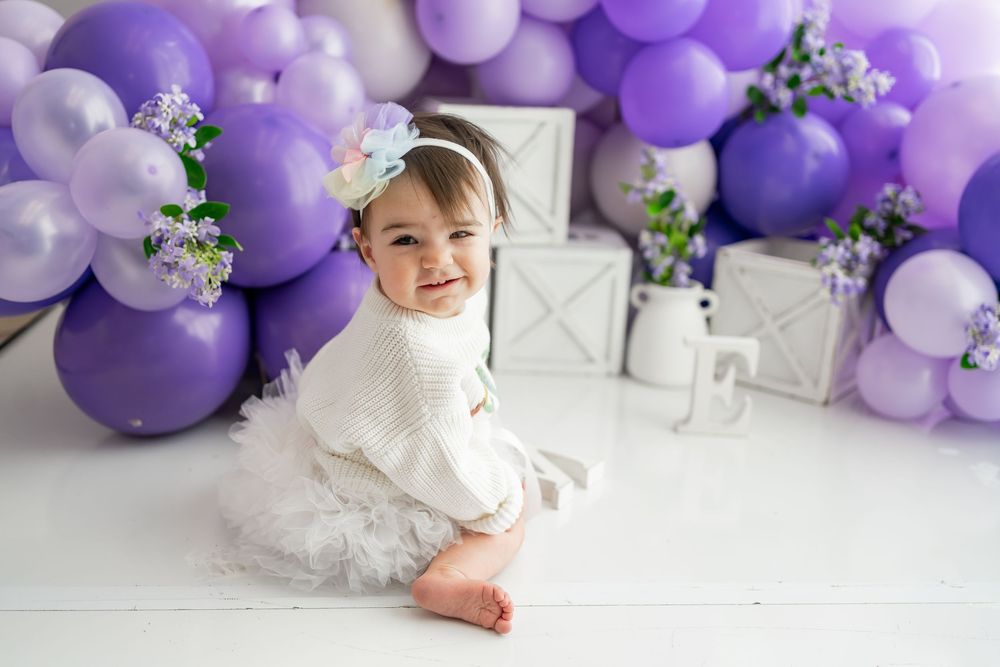Baby in white dress sits on floor with purple balloon arch and floral decor for first birthday photoshoot.