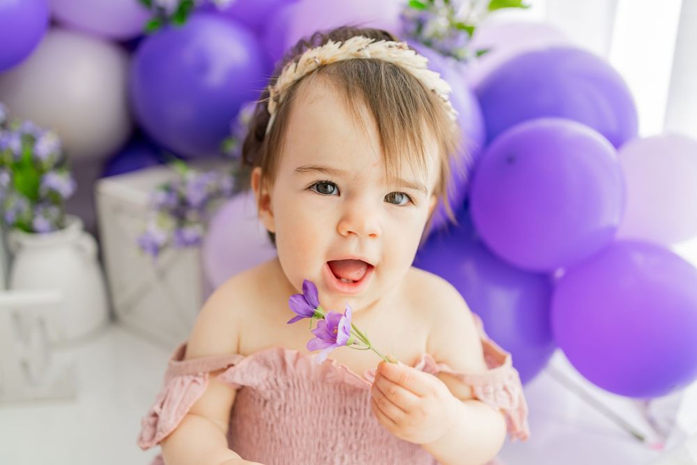 A baby wearing a pink dress sticks tongue out while posing with purple balloons and floral decorations in the background.