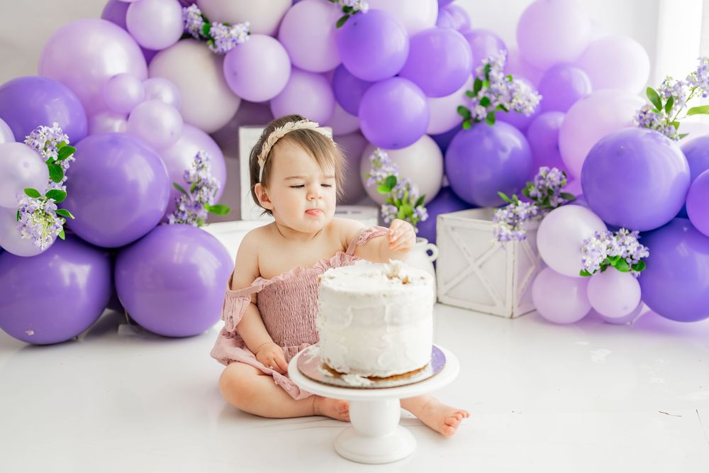 First birthday party setup with purple balloon arch and white cake on stand against white background.
