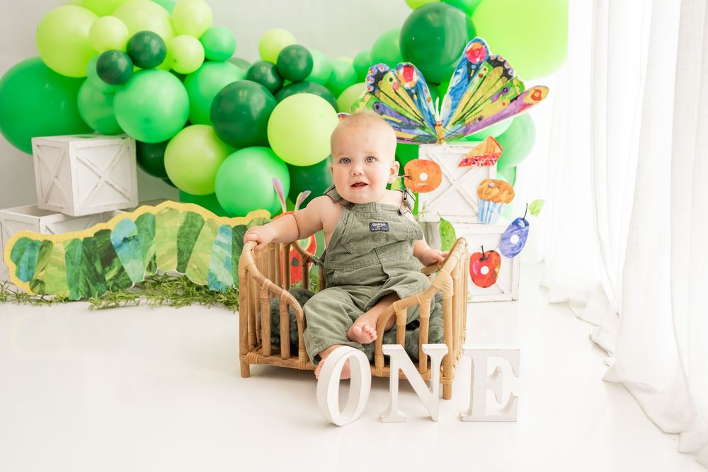 First birthday party setup with green balloons, decorations and a wooden high chair against white walls.