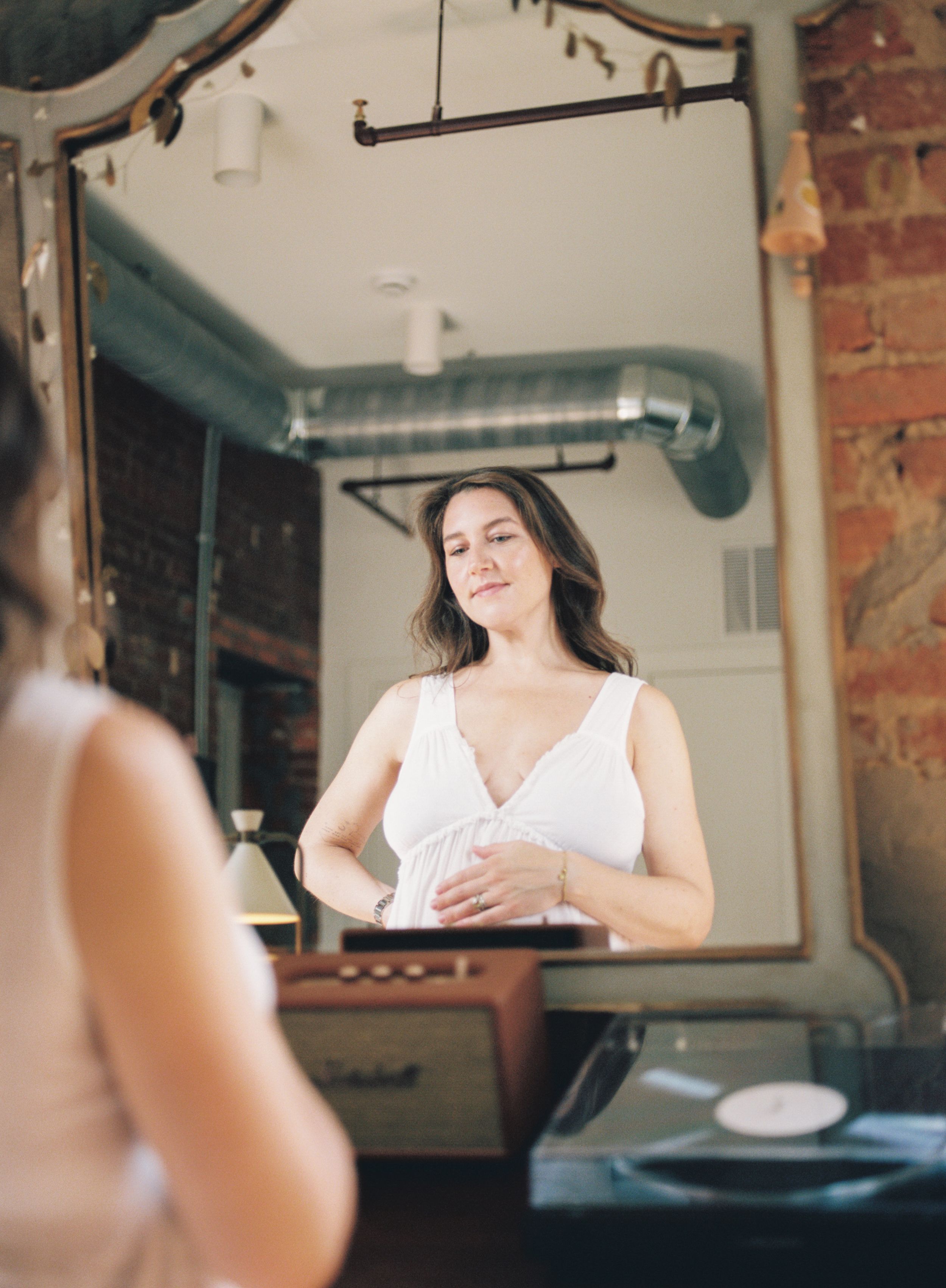 A person in a white sleeveless top looks at their reflection in a vintage-style mirror against exposed brick walls.