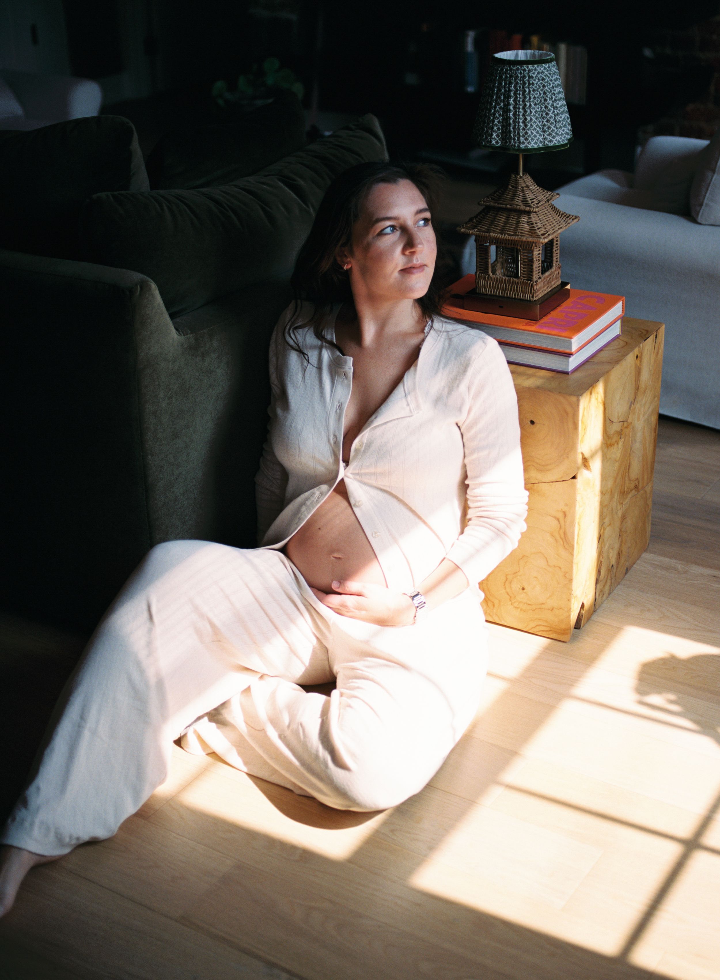 A person in a white outfit sits on the floor beside a side table with a lamp, illuminated by natural sunlight through windows.