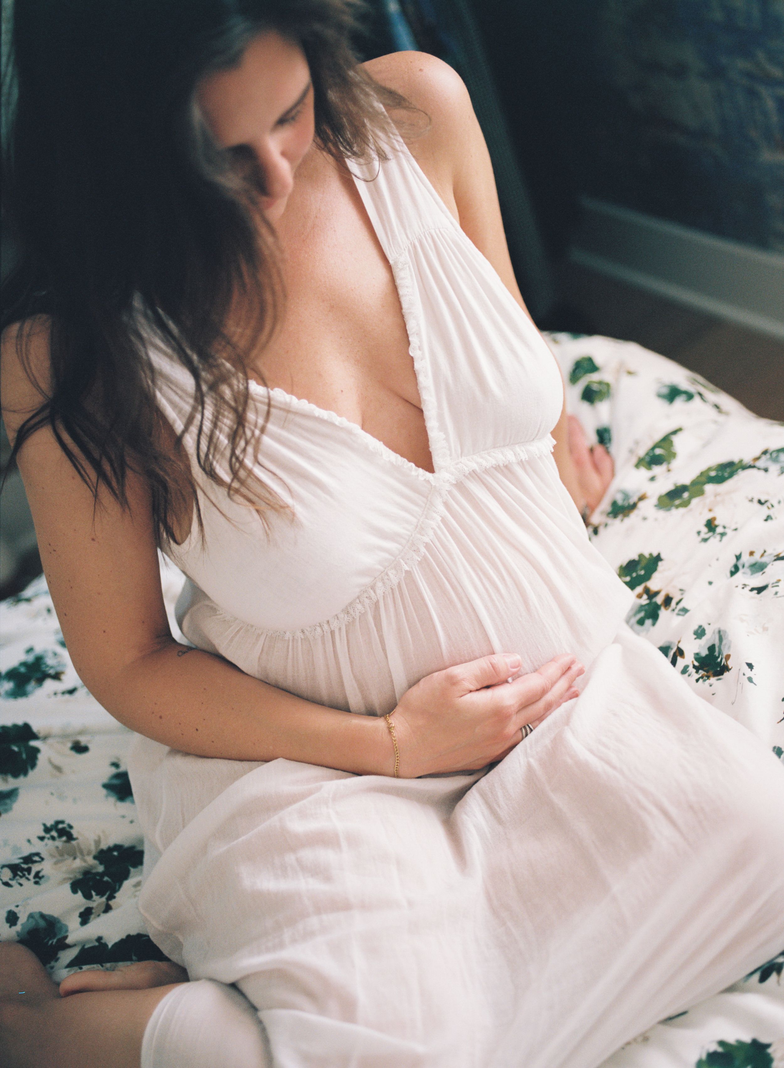 Woman in white flowing dress lounges on floral bedding in soft natural light.