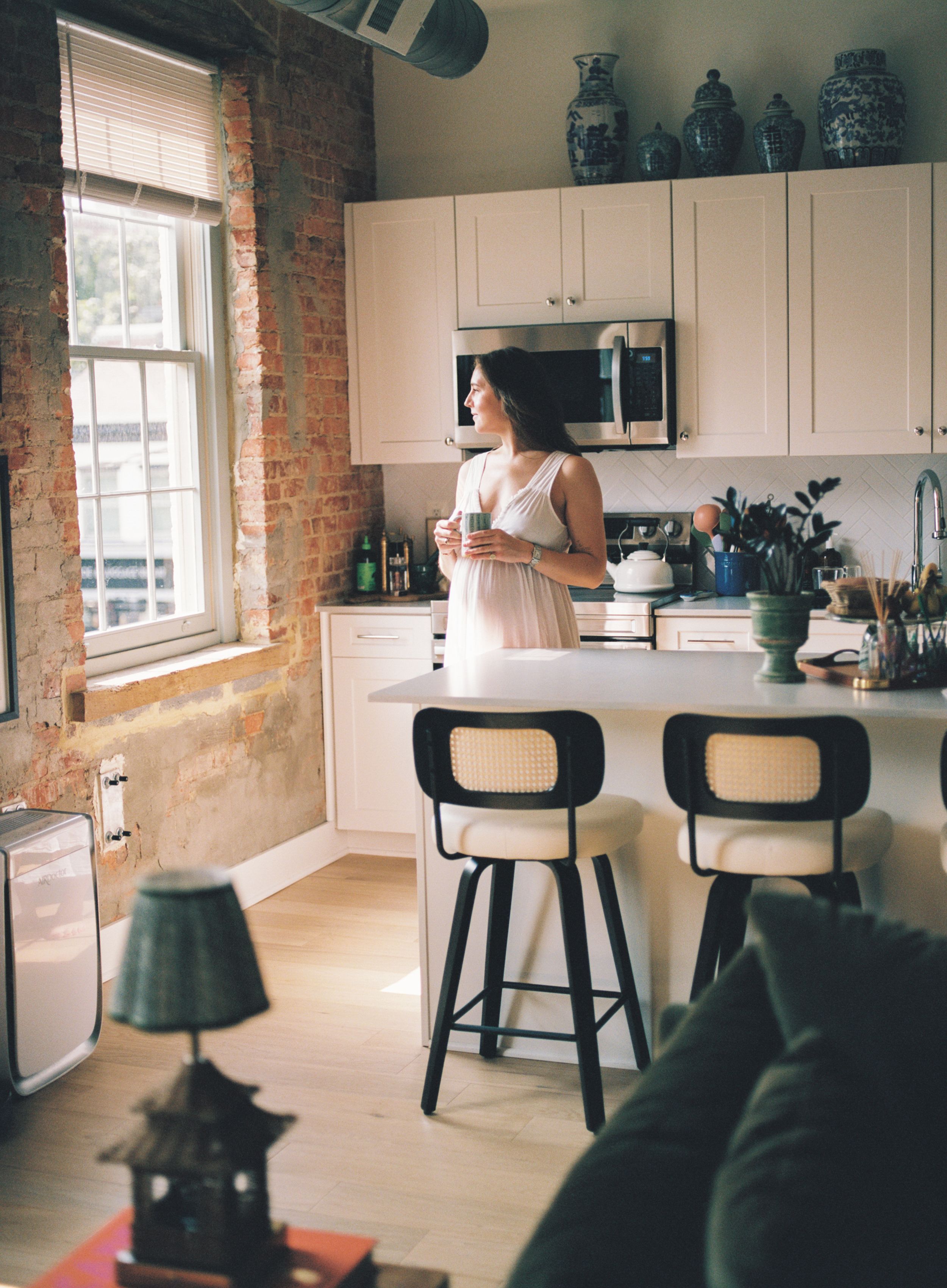 A rustic kitchen interior with exposed brick walls, white cabinets, and black counter stools in natural lighting.