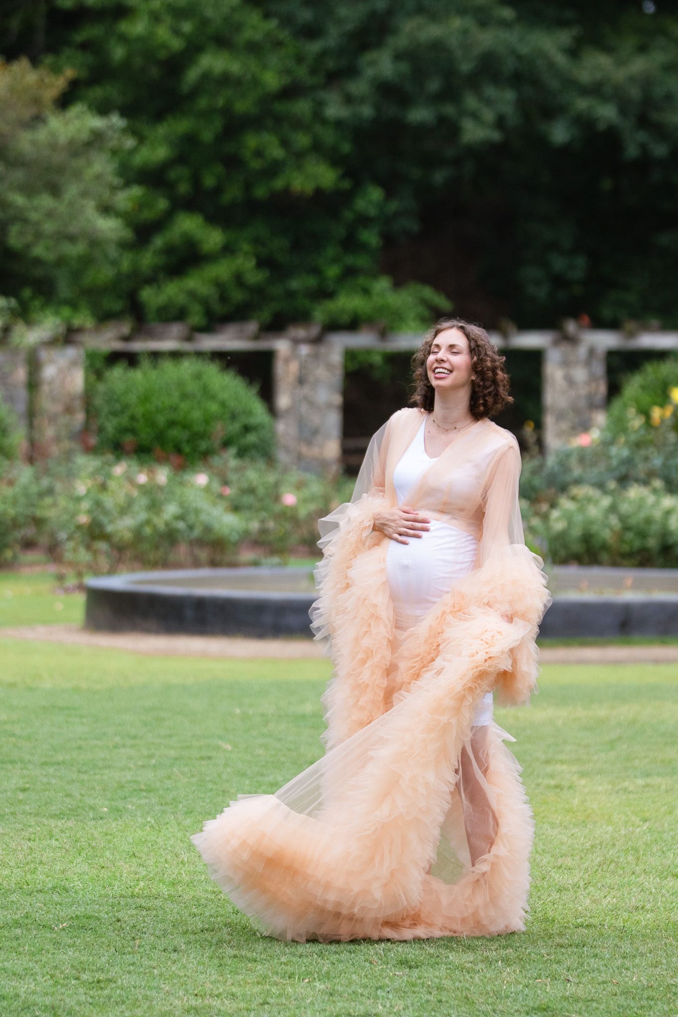 A woman in a flowing peach-colored tulle dress dances playfully on lush green grass at the Raleigh Rose Garden