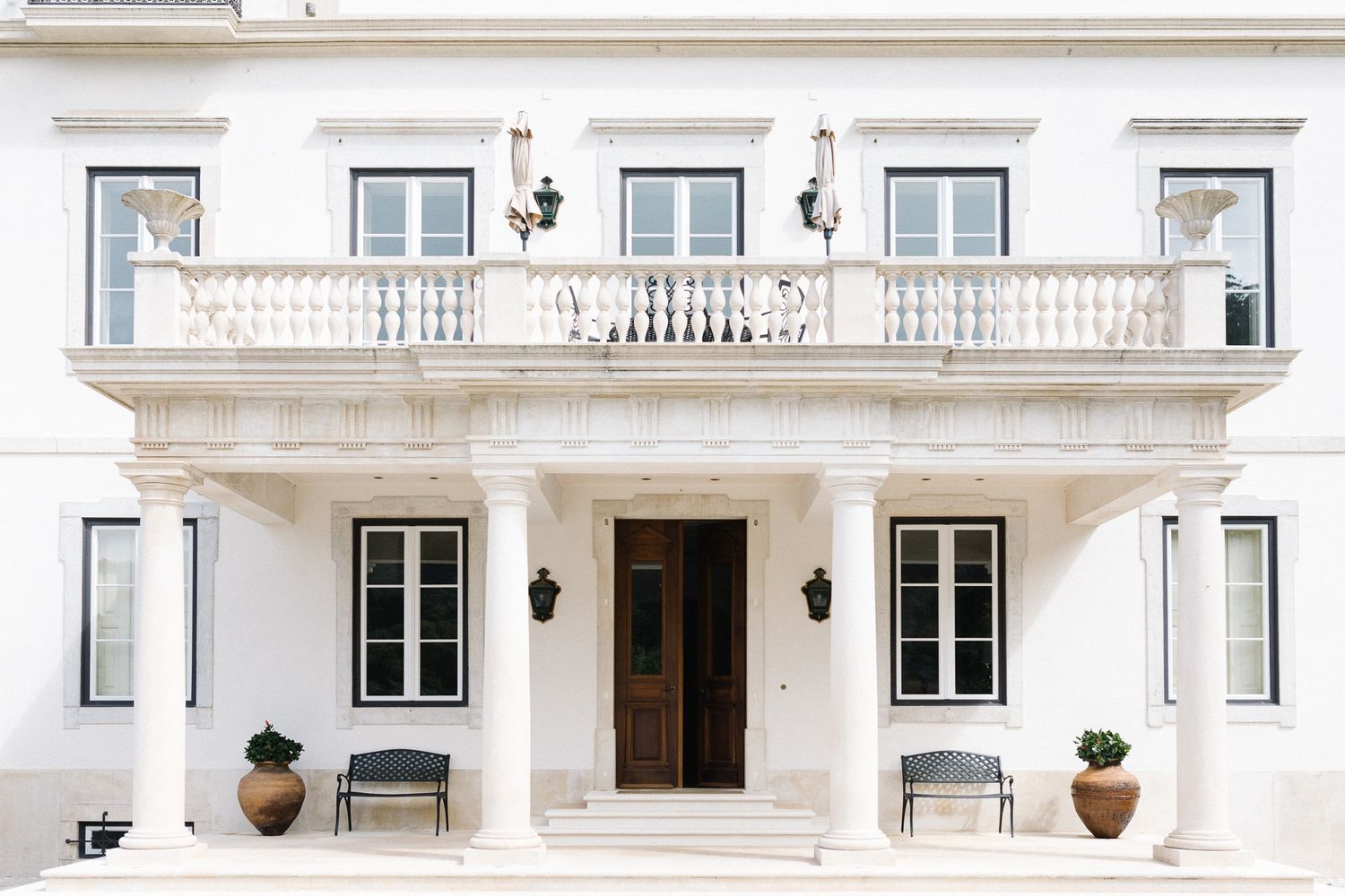 Elegant white mansion entrance with grand balcony, symmetrical windows, and decorative porch flanked by potted plants.