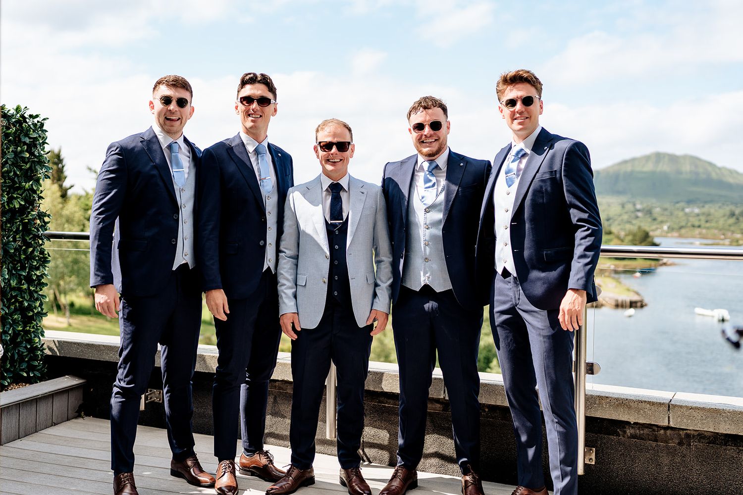 Group of groomsmen in formal suits standing together on a deck overlooking water and mountains on a sunny day.