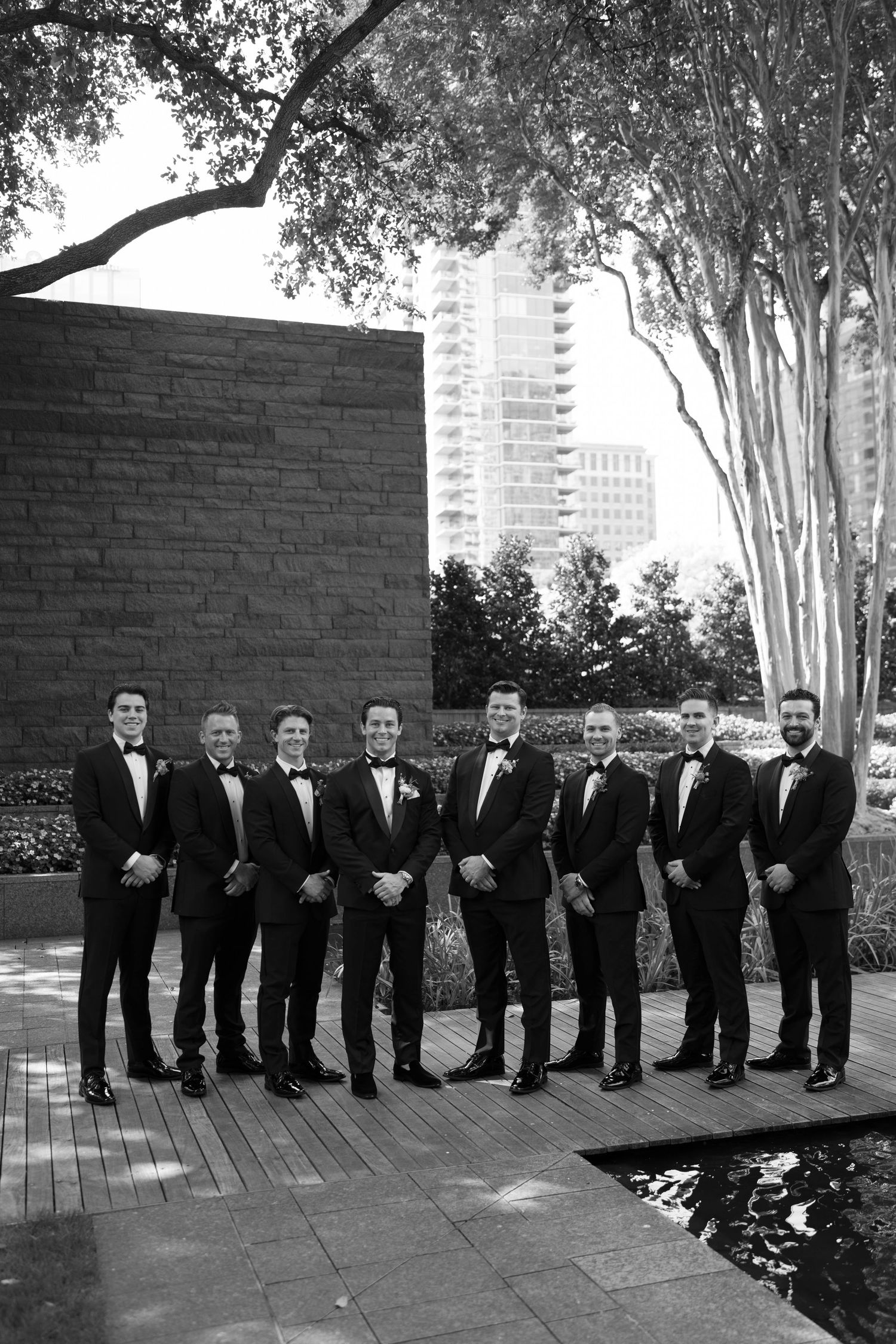Groomsmen in classic black tuxedos pose outdoors in front of brick wall and trees with city buildings in background.