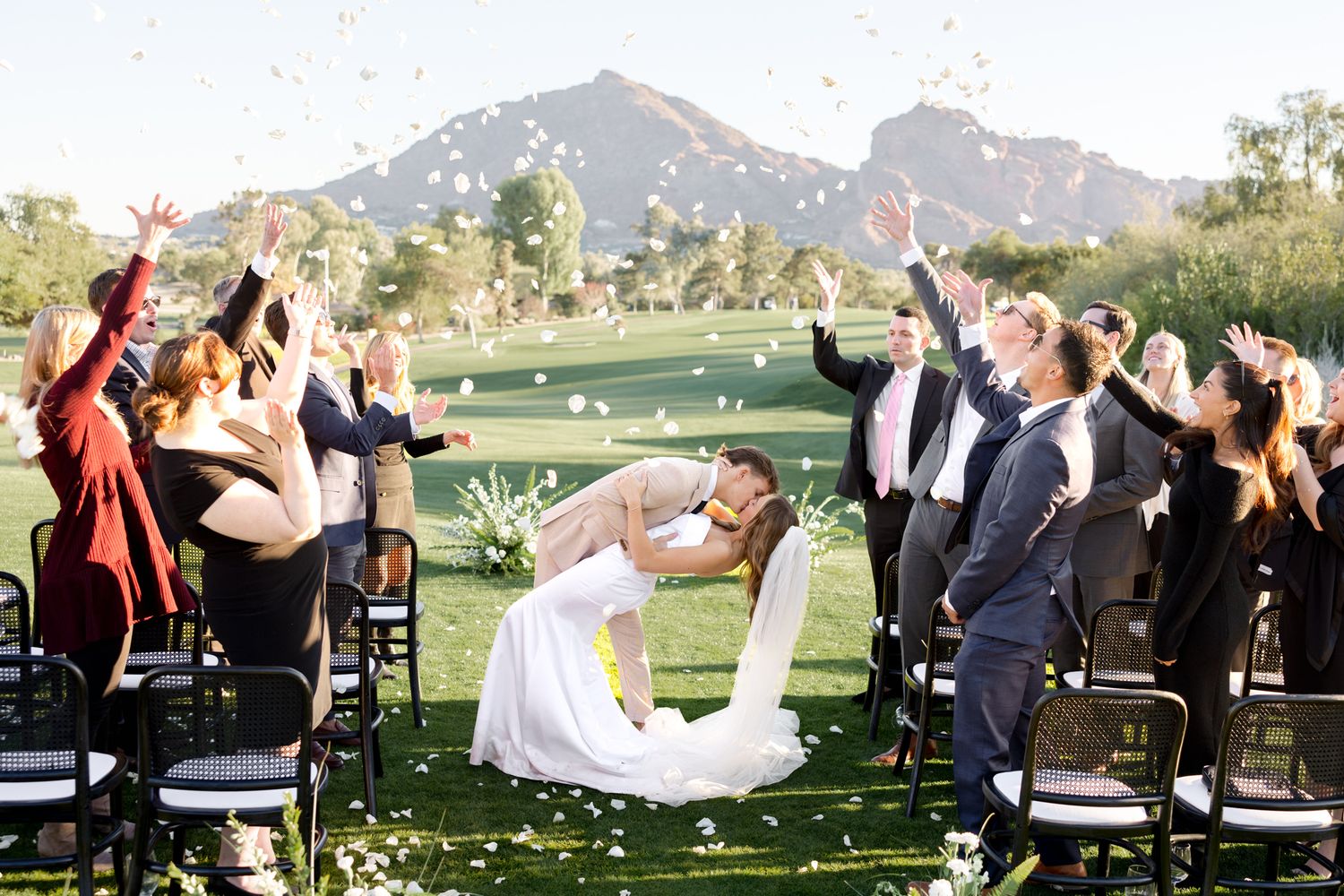 Wedding ceremony guests toss flower petals in celebration as newlyweds walk down the outdoor aisle at sunset.