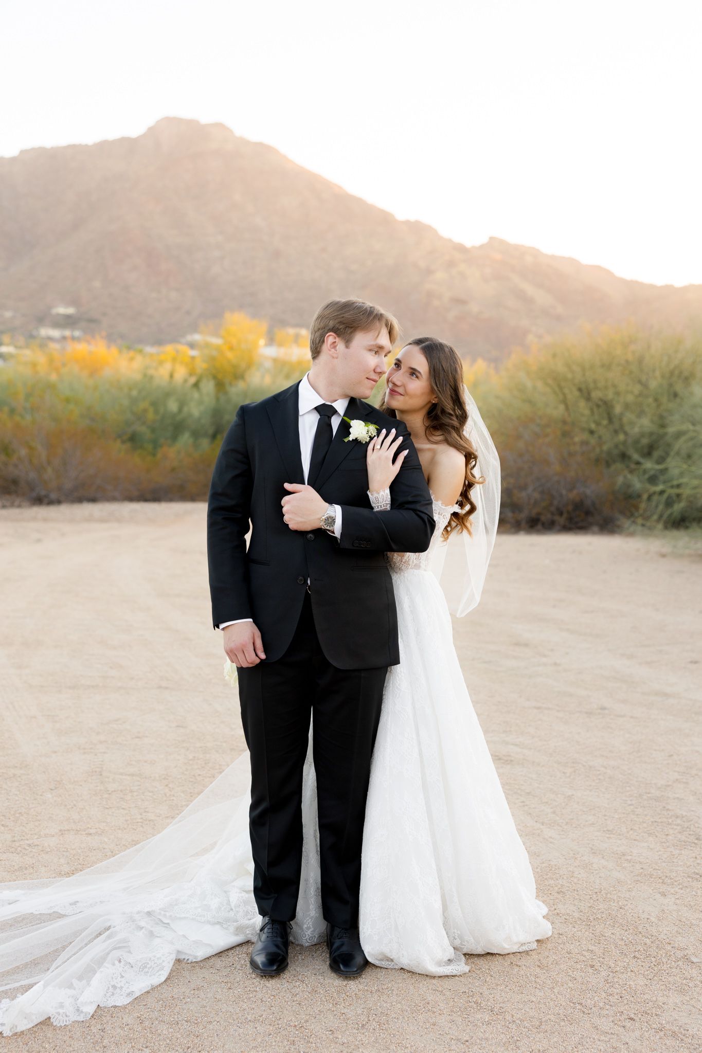 A couple shares intimate moments at sunset in a desert landscape with mountain views and a flowing white wedding dress.