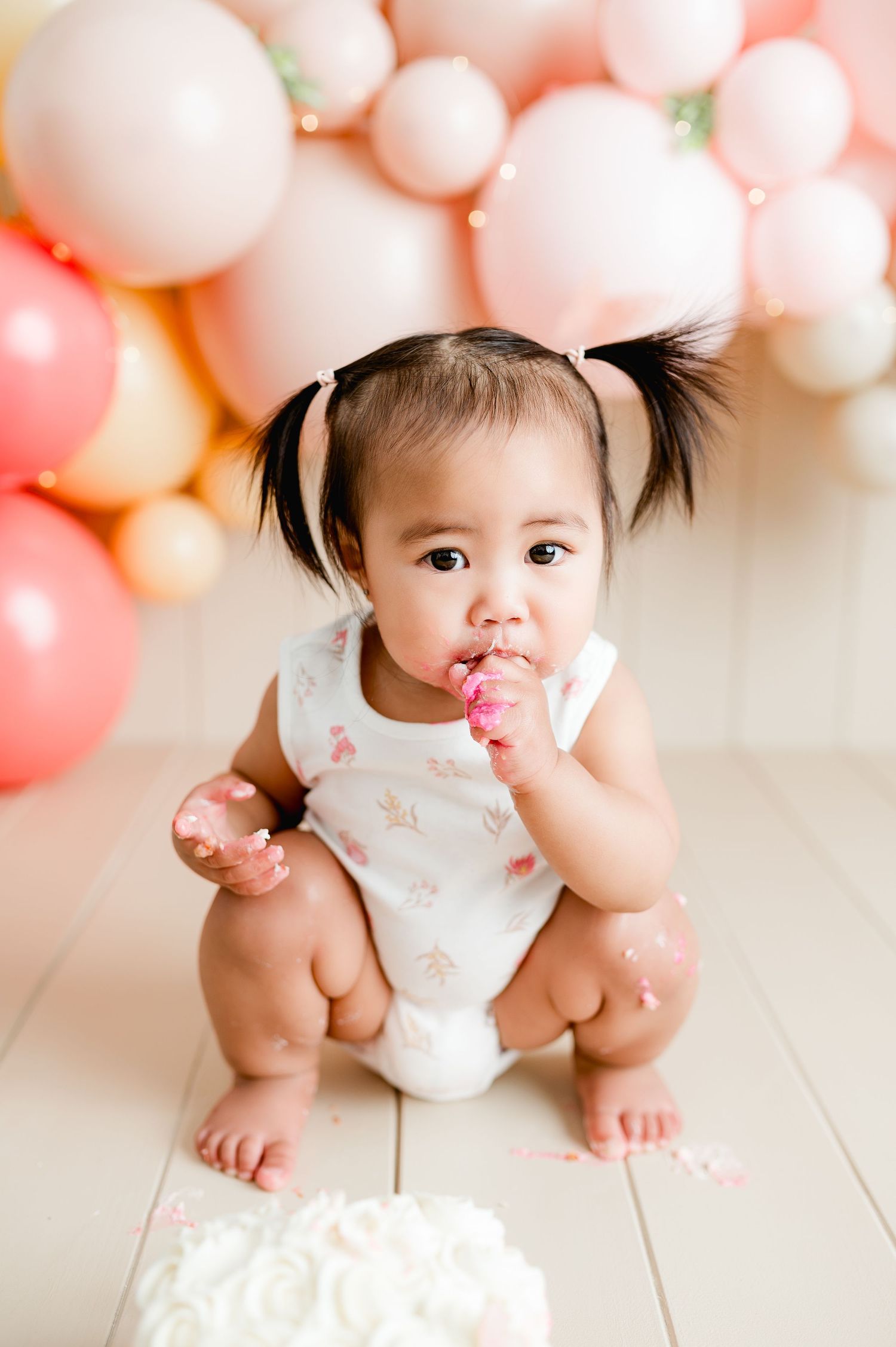 Baby in white onesie among pink and peach balloon decorations lying on white wooden floor for birthday photo shoot.