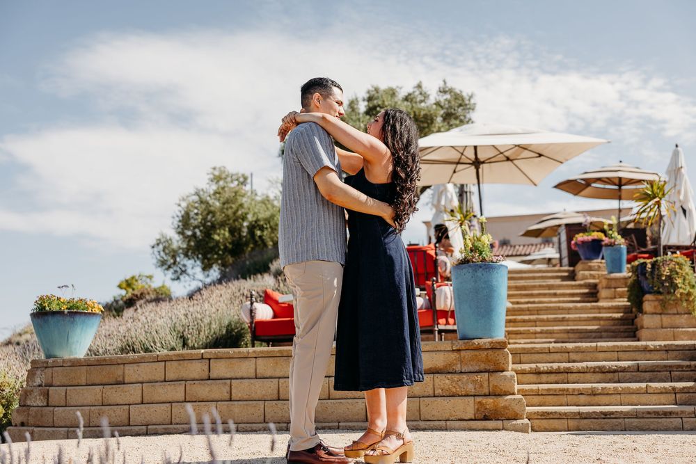 A couple embraces on outdoor stone steps with decorative patio umbrellas and potted plants in a sunny garden setting.
