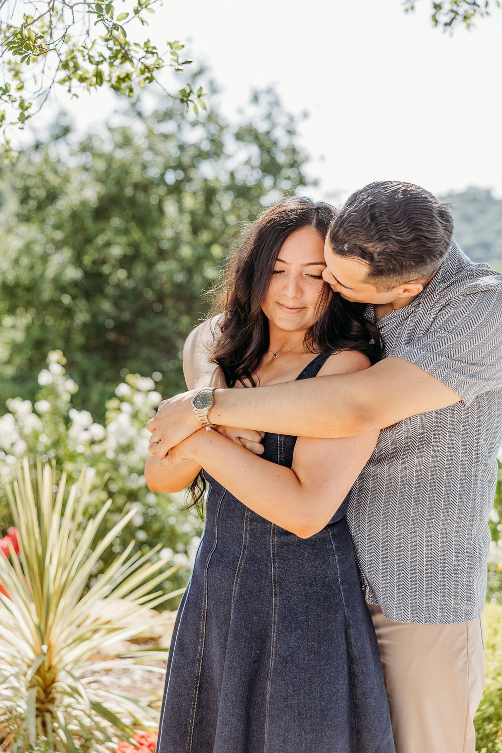 A couple in an intimate embrace outdoors with denim dress and striped shirt among green foliage.