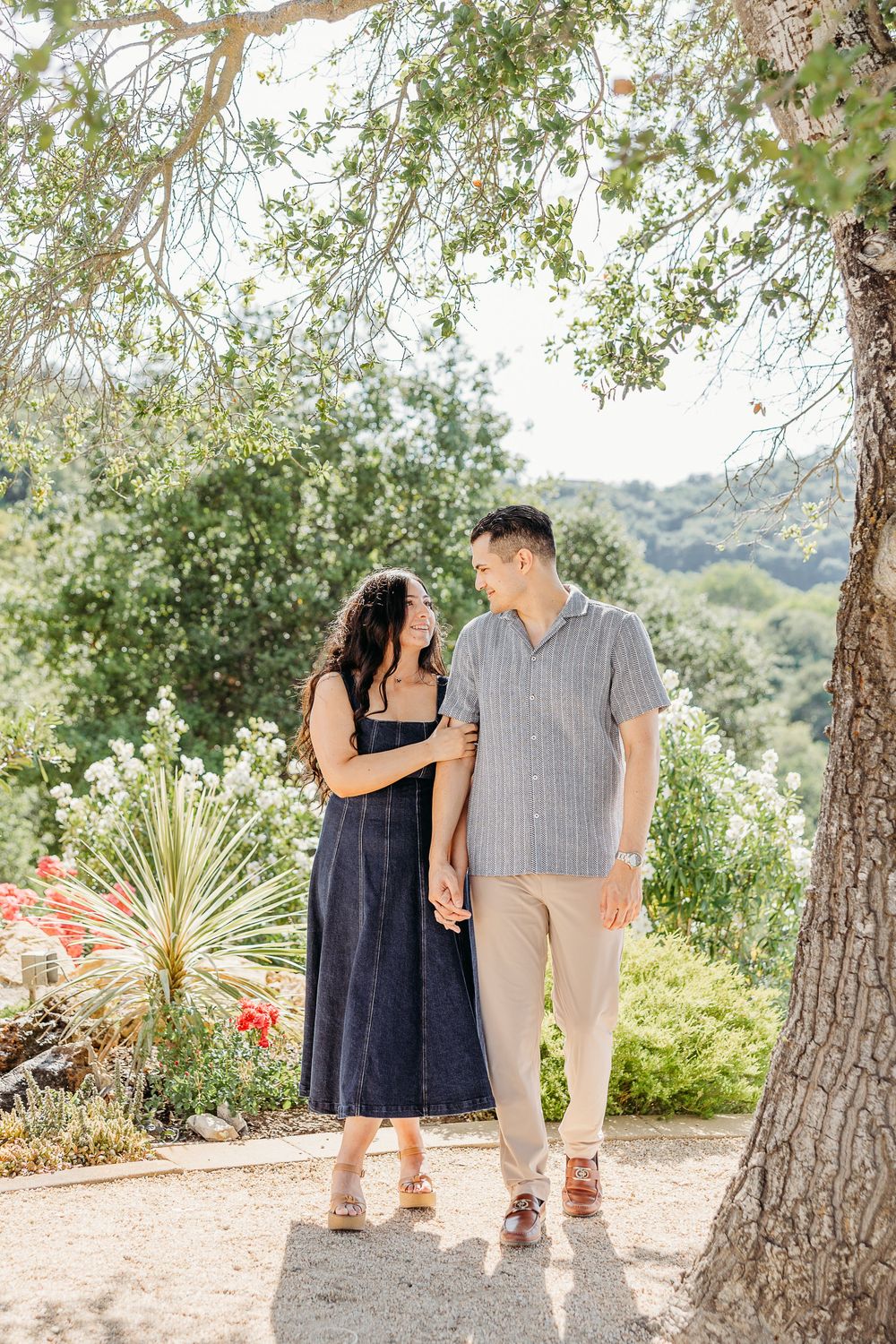 Two people walking together on a scenic path surrounded by trees and desert plants in a natural outdoor setting.