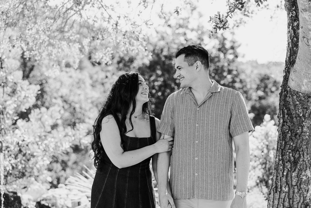 A romantic couple poses together under shady trees in a natural outdoor setting.