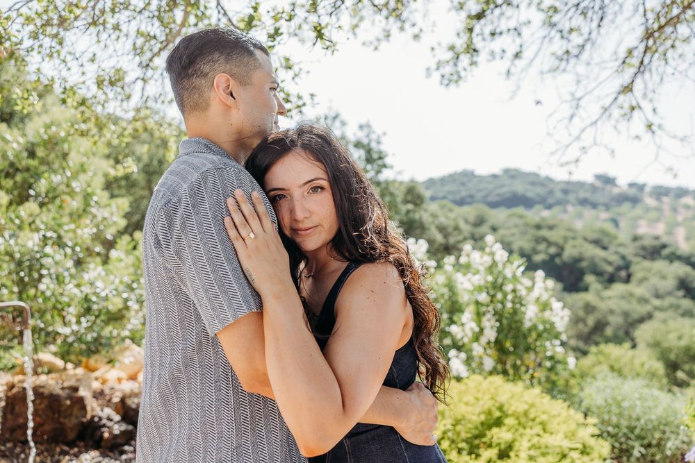 Two people share a tender embrace while standing in a sun-dappled garden with green hills in the background.