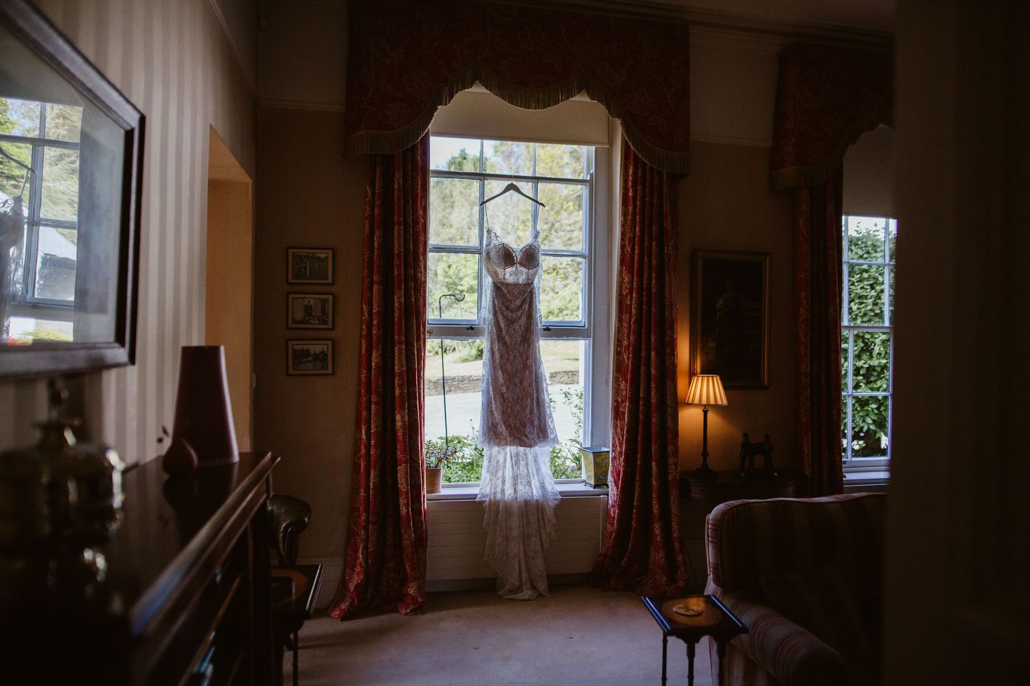 A vintage pink wedding dress hangs in a window frame, backlit by natural daylight in a cozy room with red curtains.