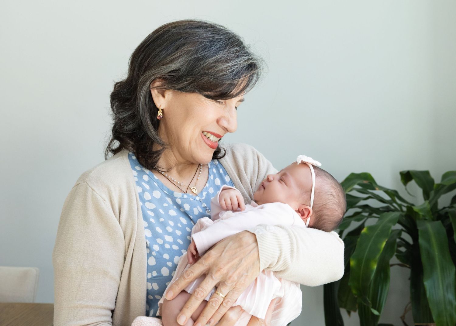 A person wearing a beige cardigan and blue polka dot dress holds a sleeping newborn baby near a houseplant.