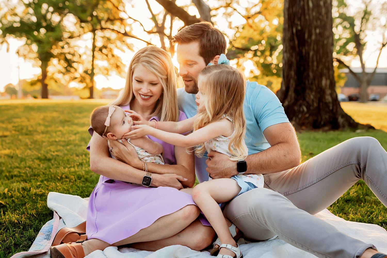 A family laughs and plays together while relaxing on a blanket in a sunlit park during golden hour.