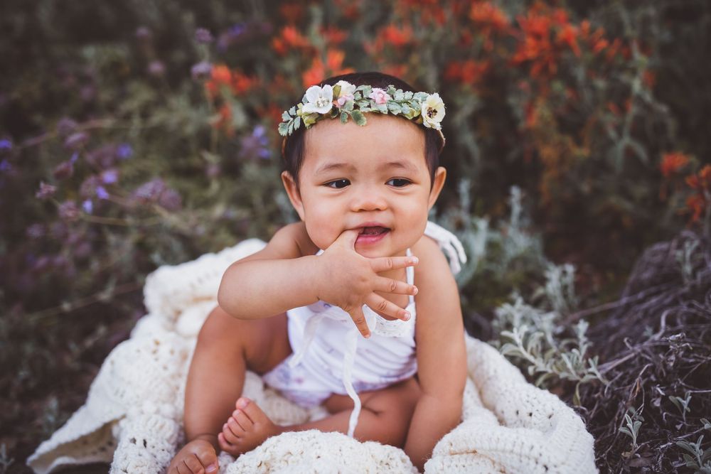 A baby wearing a white lace dress sits outdoors against a natural background during a photo session.