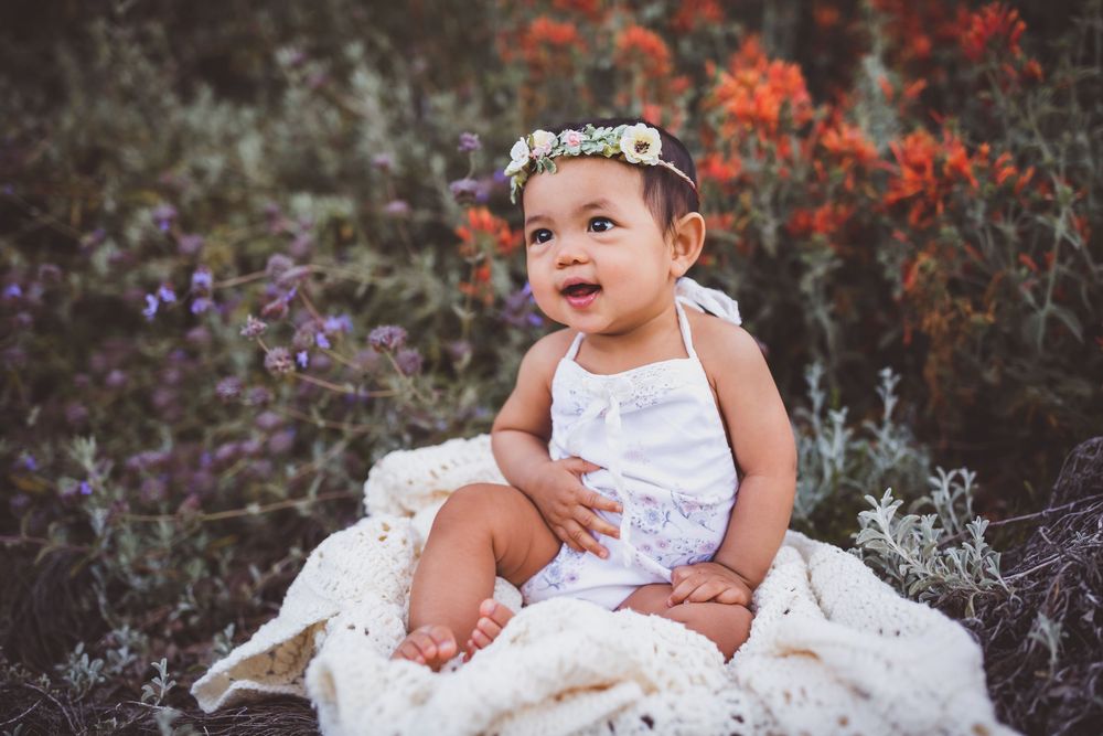 Baby sitting in white lace dress surrounded by colorful wildflowers and plants in outdoor garden setting.