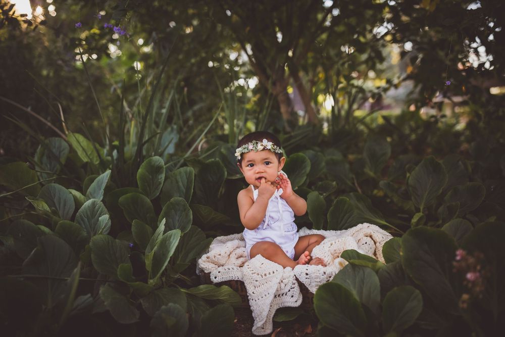 A small child in a white dress sits among lush green foliage at sunset wearing a floral crown.
