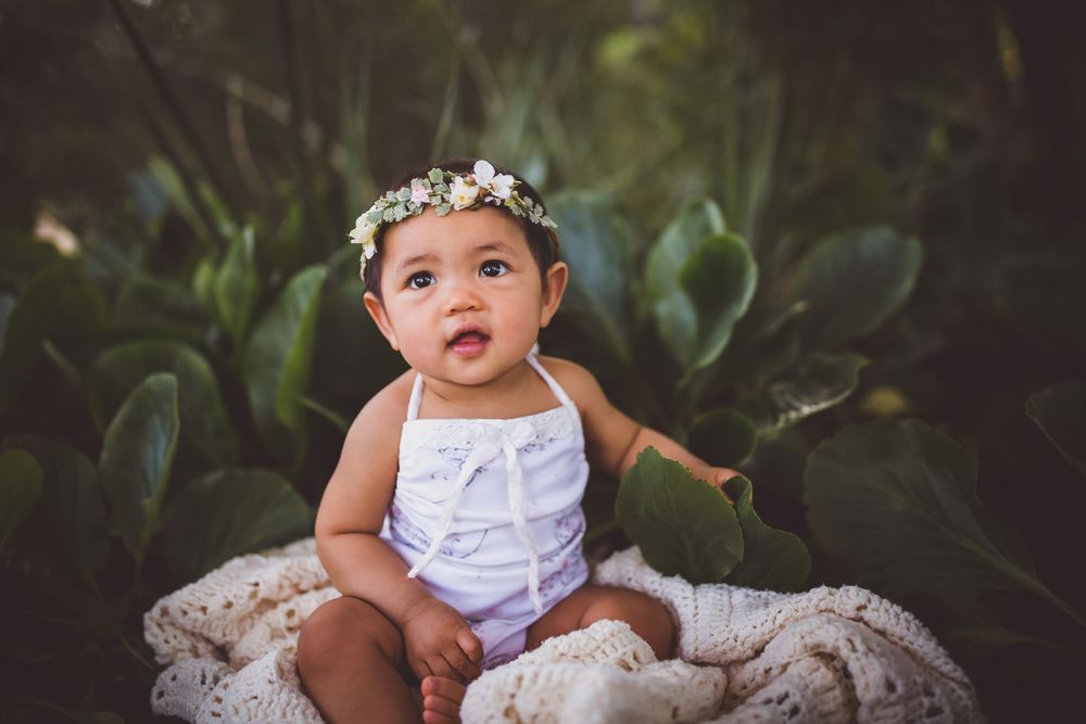 A baby in white clothing and floral headpiece sits on textured fabric surrounded by green plants.