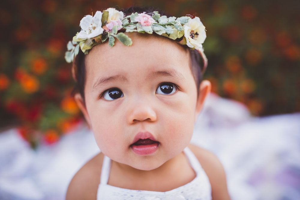 Baby wearing white dress and delicate floral crown gazes at camera against colorful bokeh background.