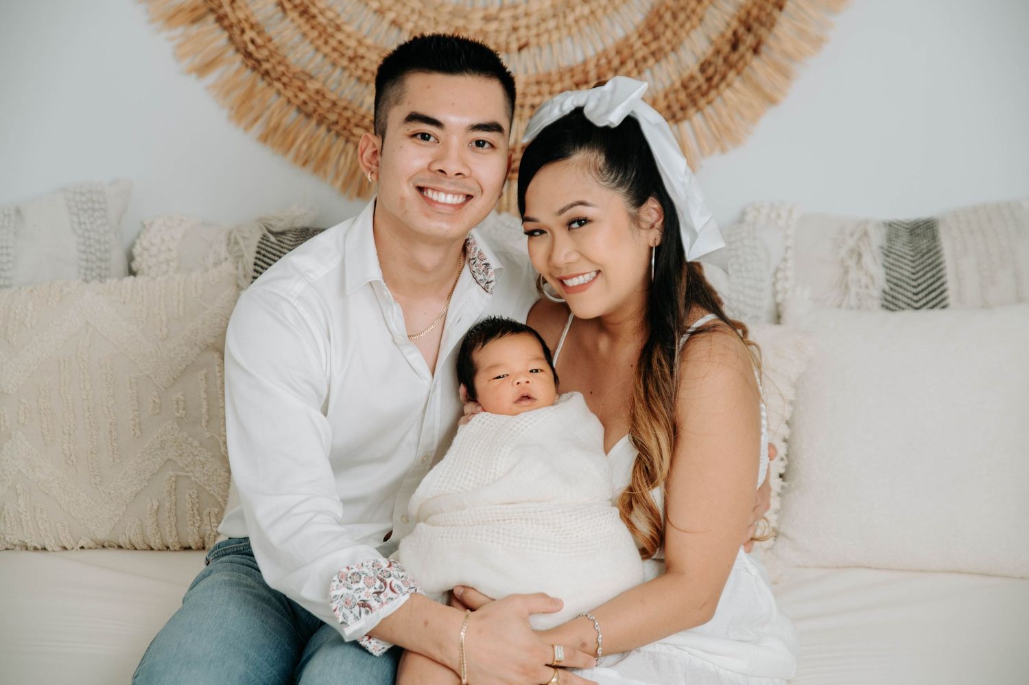 A loving family poses together on a cream couch with a decorative fan wall hanging behind them.