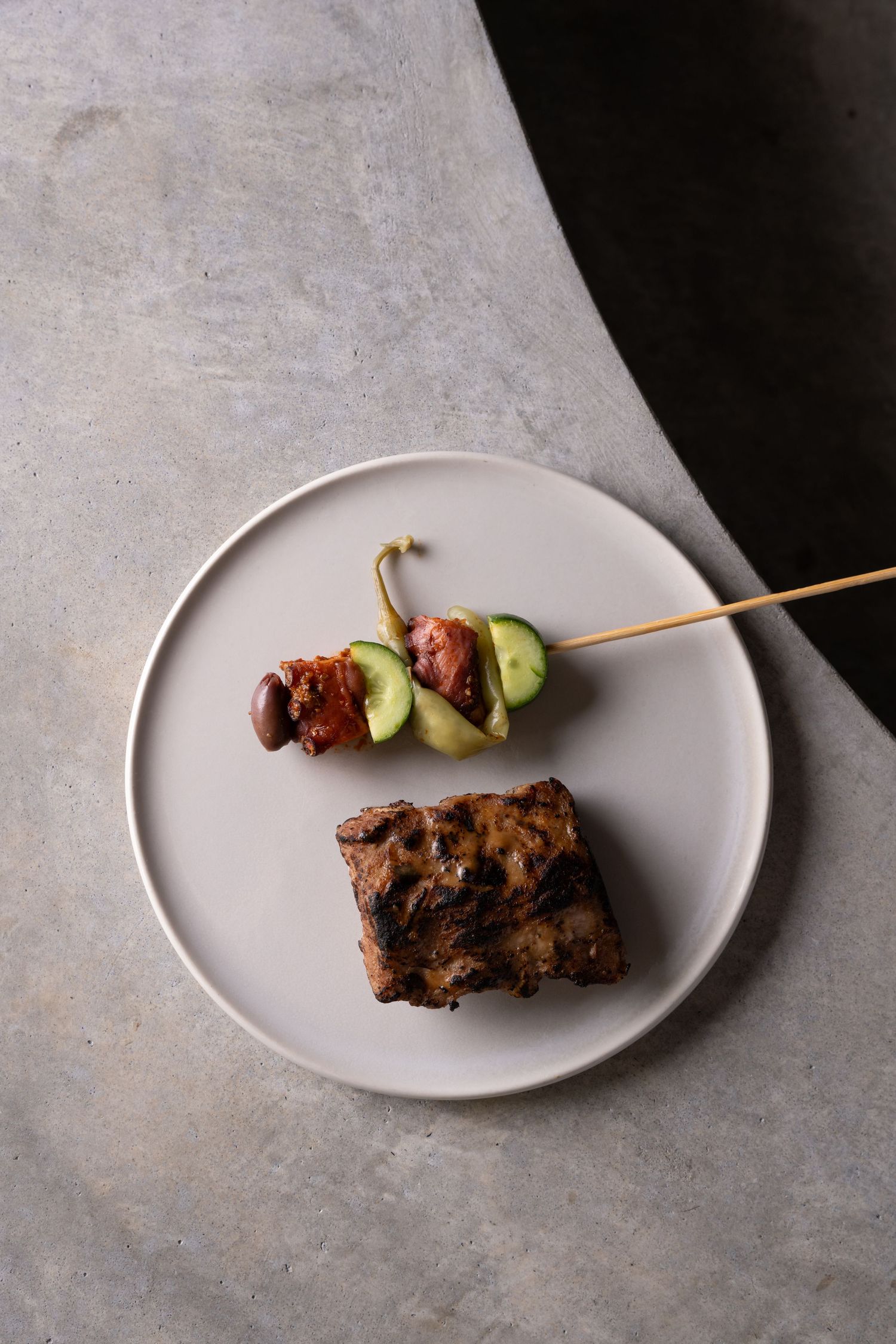 Minimalist plate featuring a grilled meat skewer with avocado and a rectangular piece of grilled meat, styled on a concrete surface.