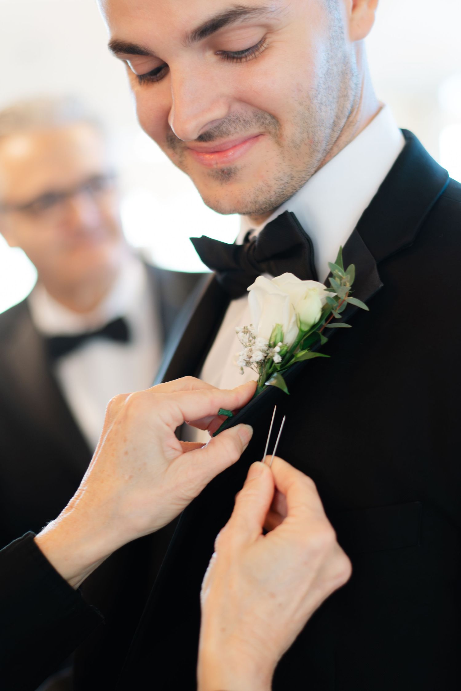 Hands carefully pinning a white floral boutonniere onto the lapel of a black tuxedo jacket at a wedding in NYC.