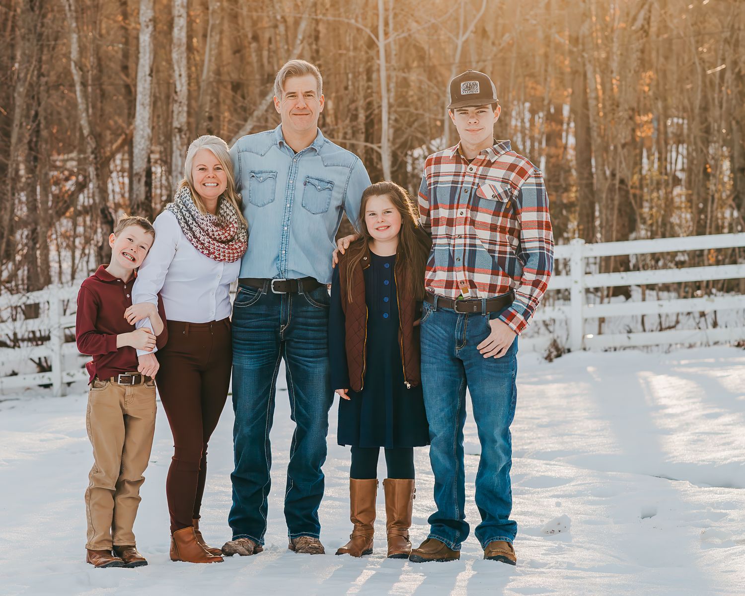 Family of five standing together on a snowy path by a white fence in winter clothing during golden hour