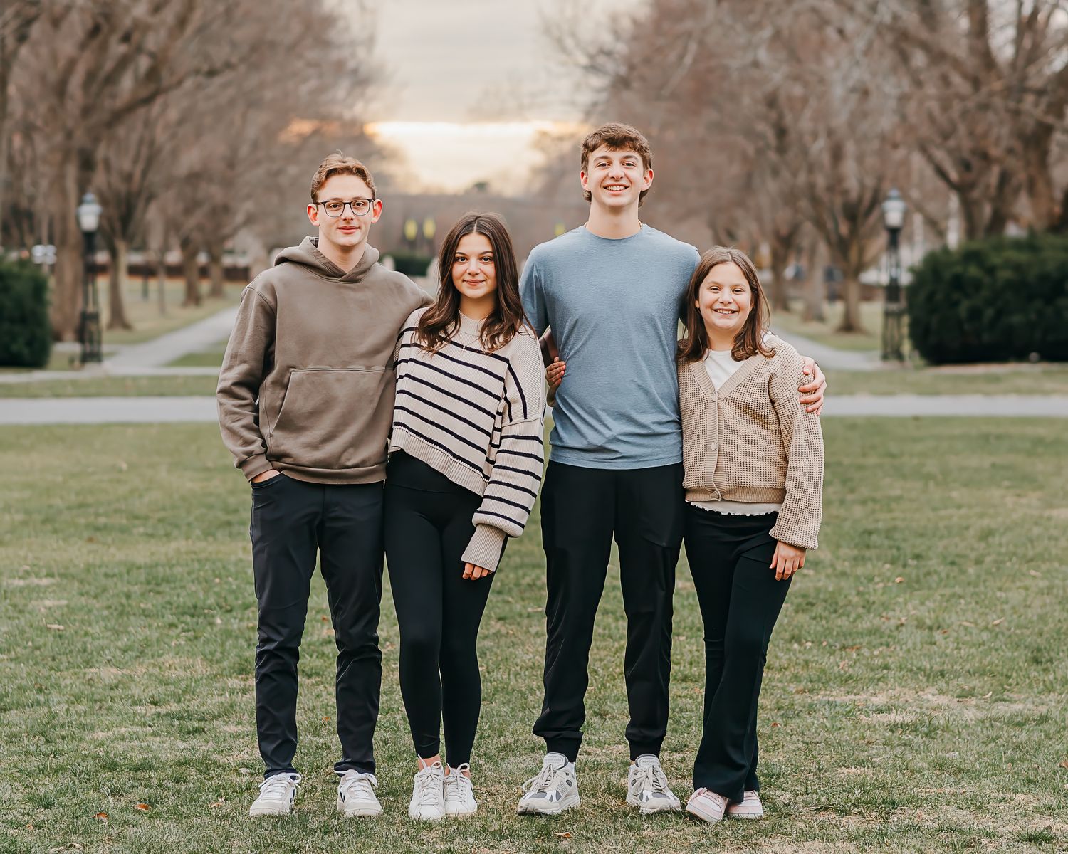 Four teenage siblings in casual sweaters and pants stand together on a grassy lawn during sunset