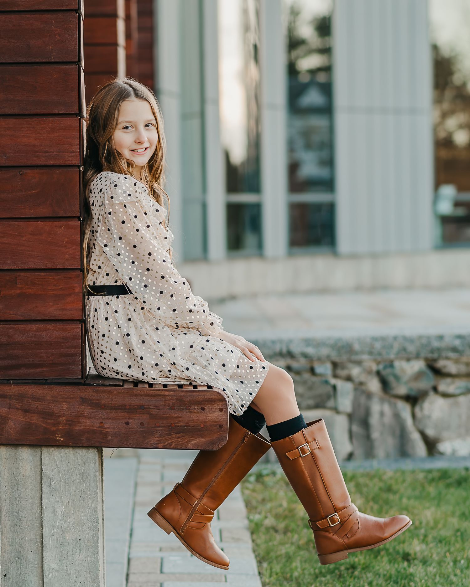 young girl in polka dot dress and brown boots sits on wooden ledge near stone wall