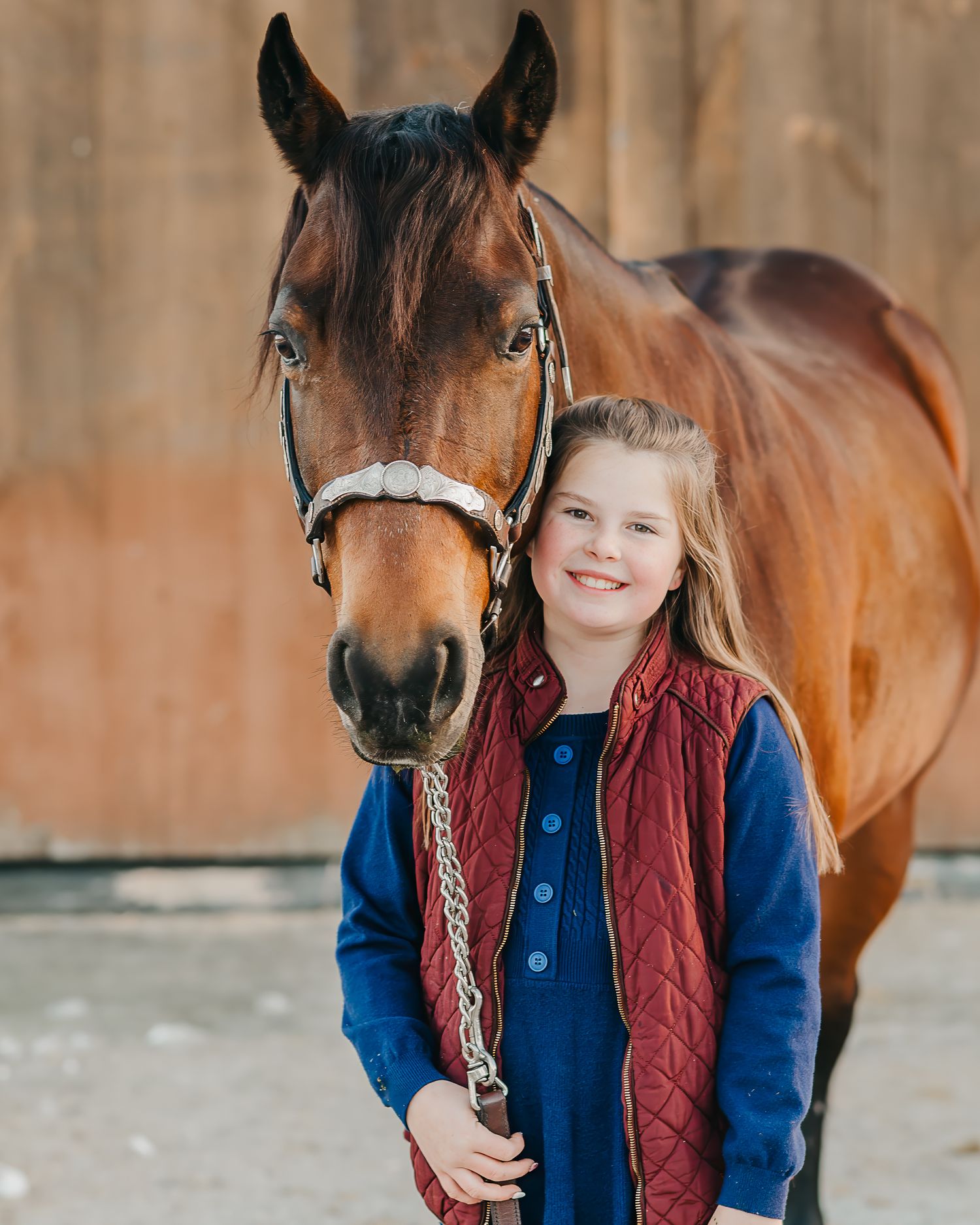 Young person in blue jacket and red vest standing with brown horse near barn.