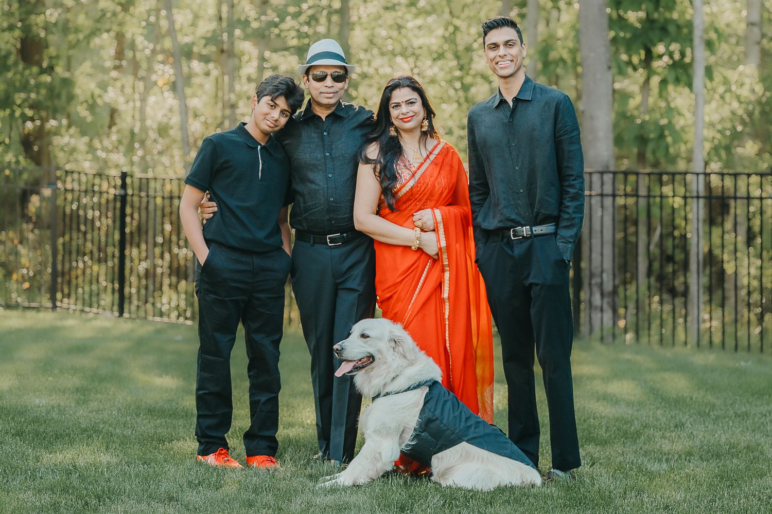 Family members wearing dark clothing pose with someone in a bright orange dress on a grassy lawn near trees and fencing.