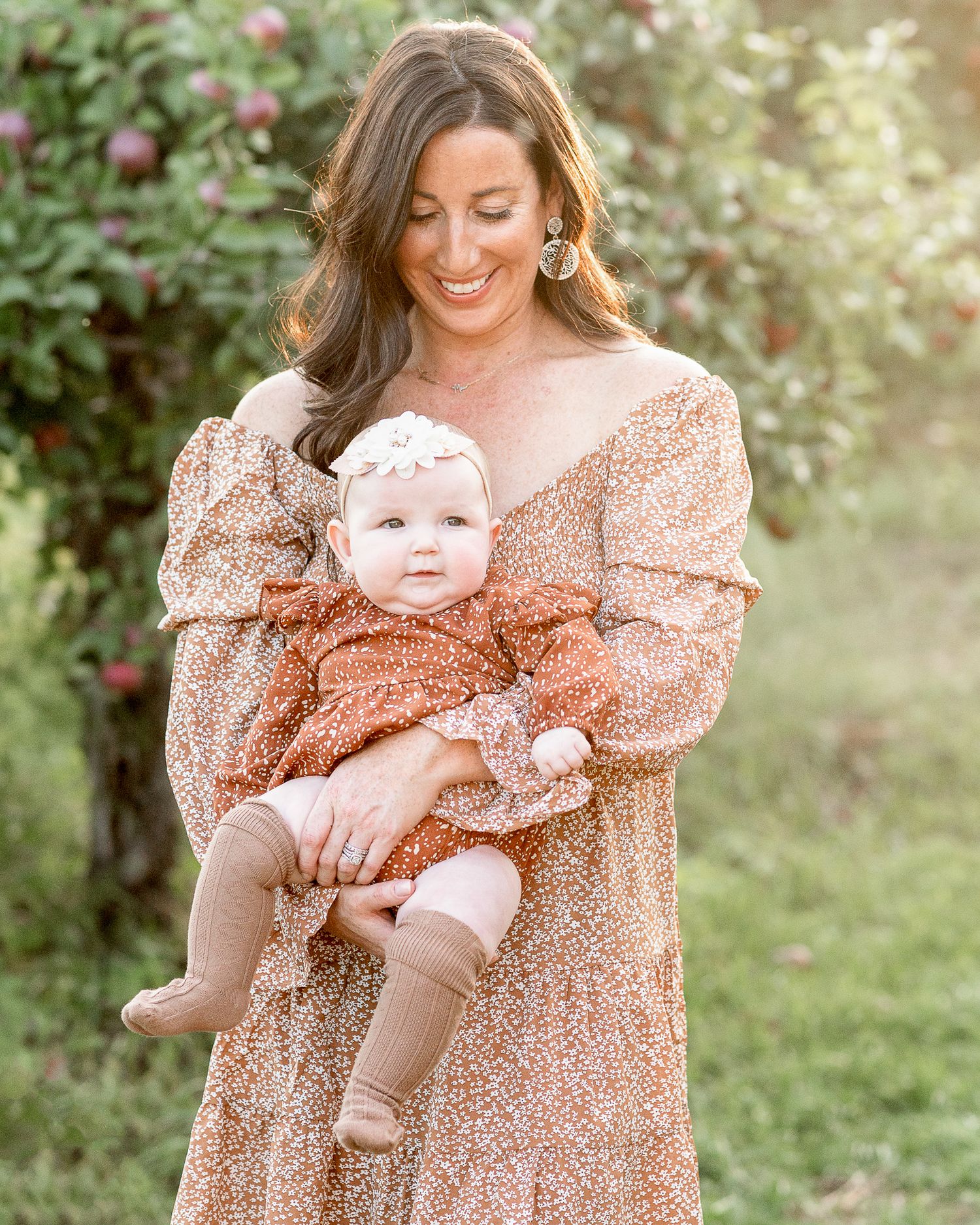 Mother and baby wearing coordinating floral print dresses pose together in a sunny garden setting in an apple orchard
