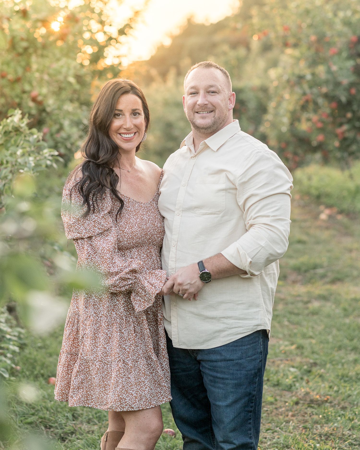 A couple poses together in an orchard during golden hour sunset wearing complementary casual outfits