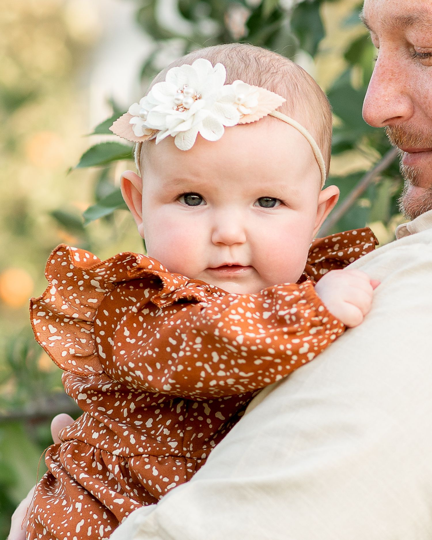 Baby wearing white floral headband and brown polka dot outfit being held in outdoor garden setting