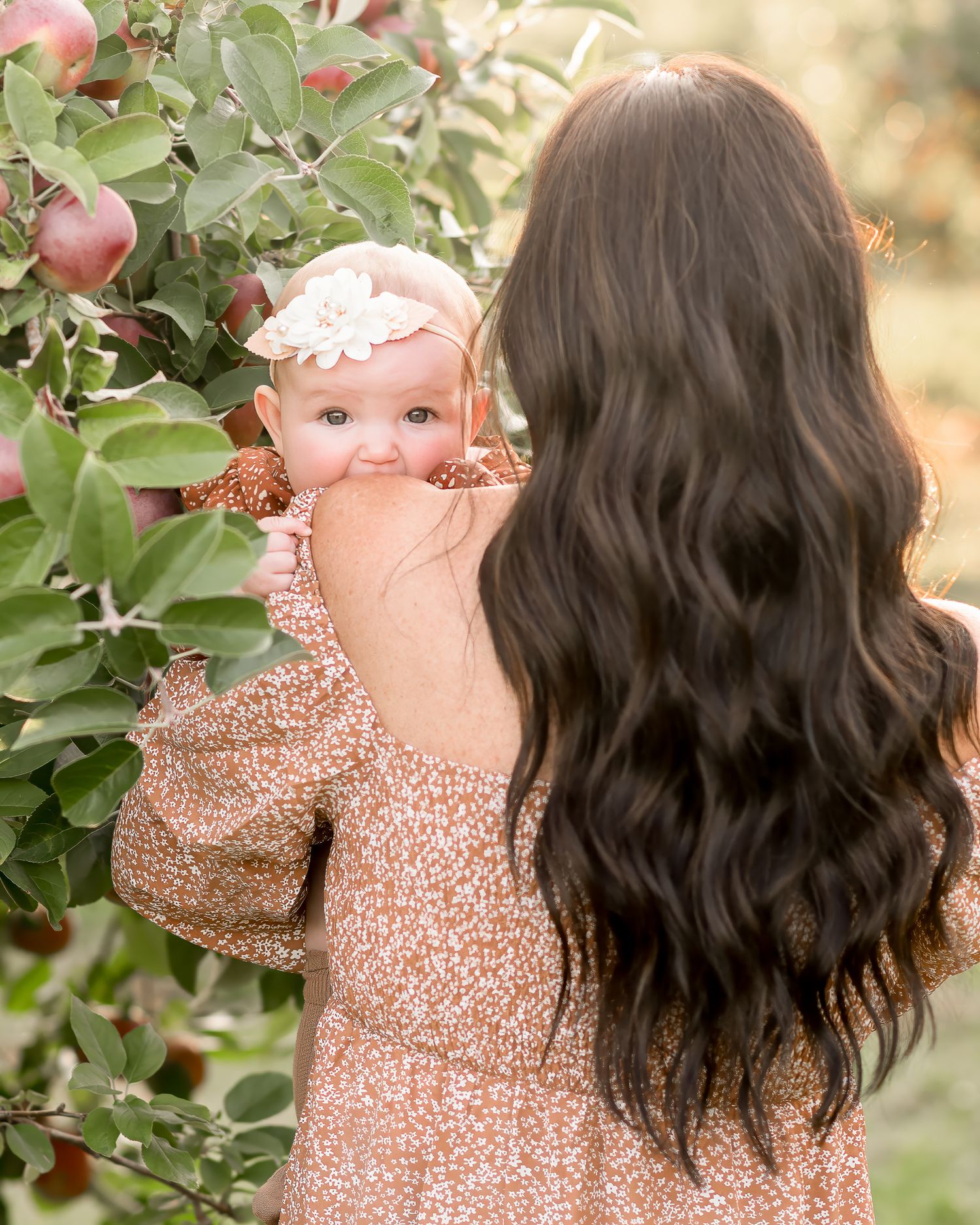 A woman in a floral dress holds a baby near an apple tree in a dreamy autumn orchard setting.