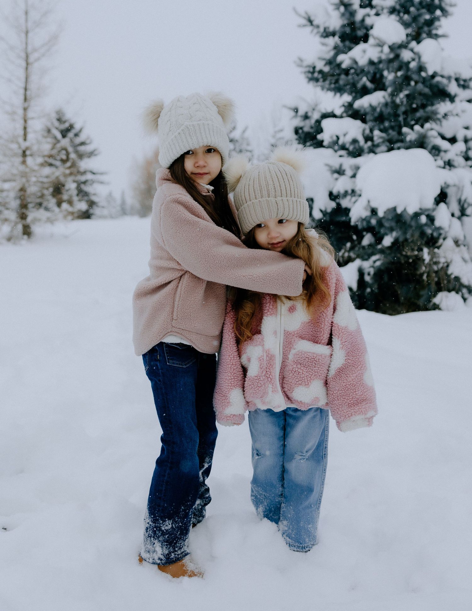 Two people in winter coats and pom-pom hats embrace while standing in snow-covered landscape.