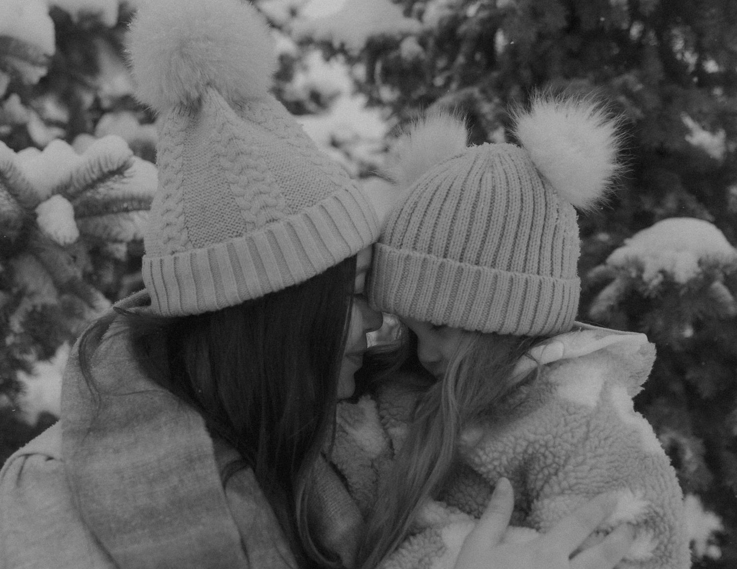 A monochrome image of two people in knit hats sharing a tender moment among snowy trees.