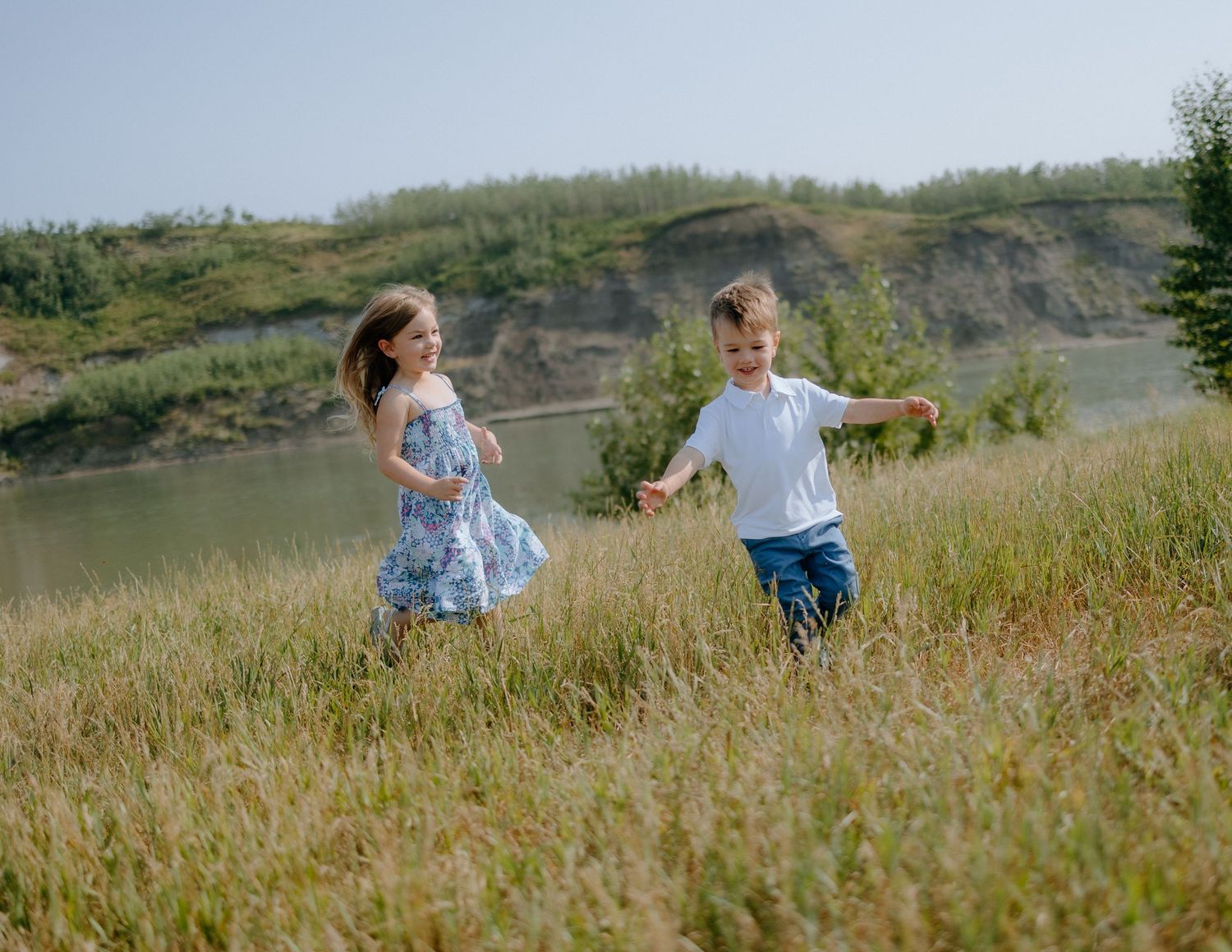 Two children running through tall grass near a cliffside lake on a sunny day.