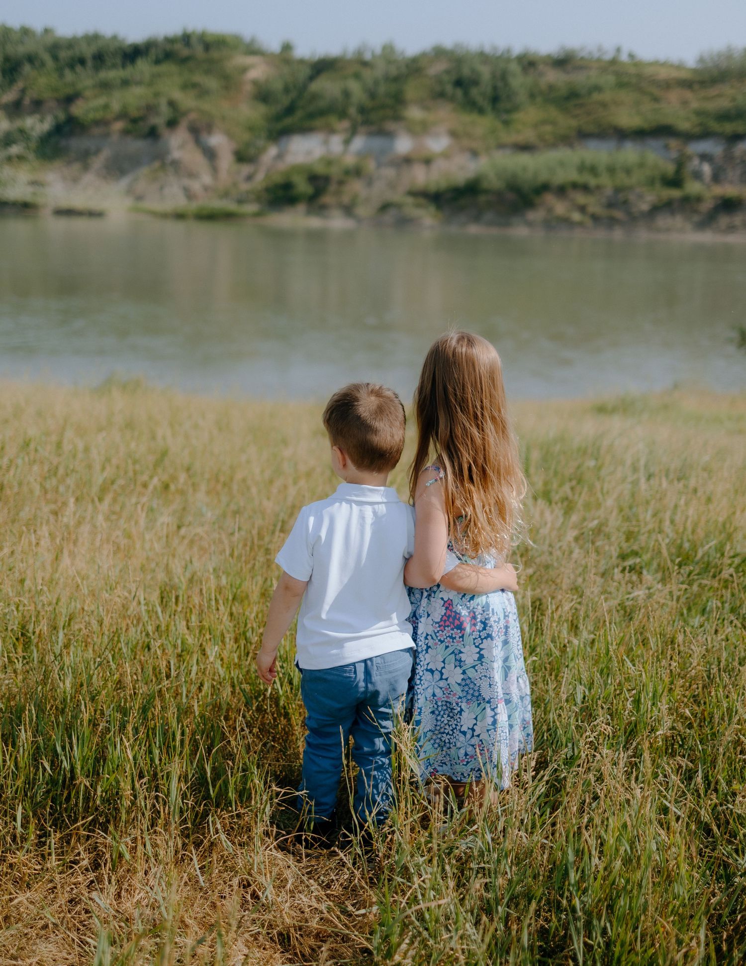 Two figures standing together overlooking a serene lake surrounded by grassy hills.