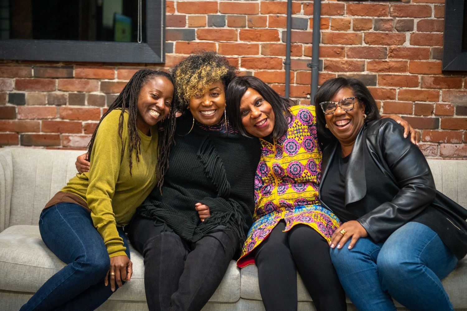Four friends sitting close together on couch against brick wall laughing and smiling.