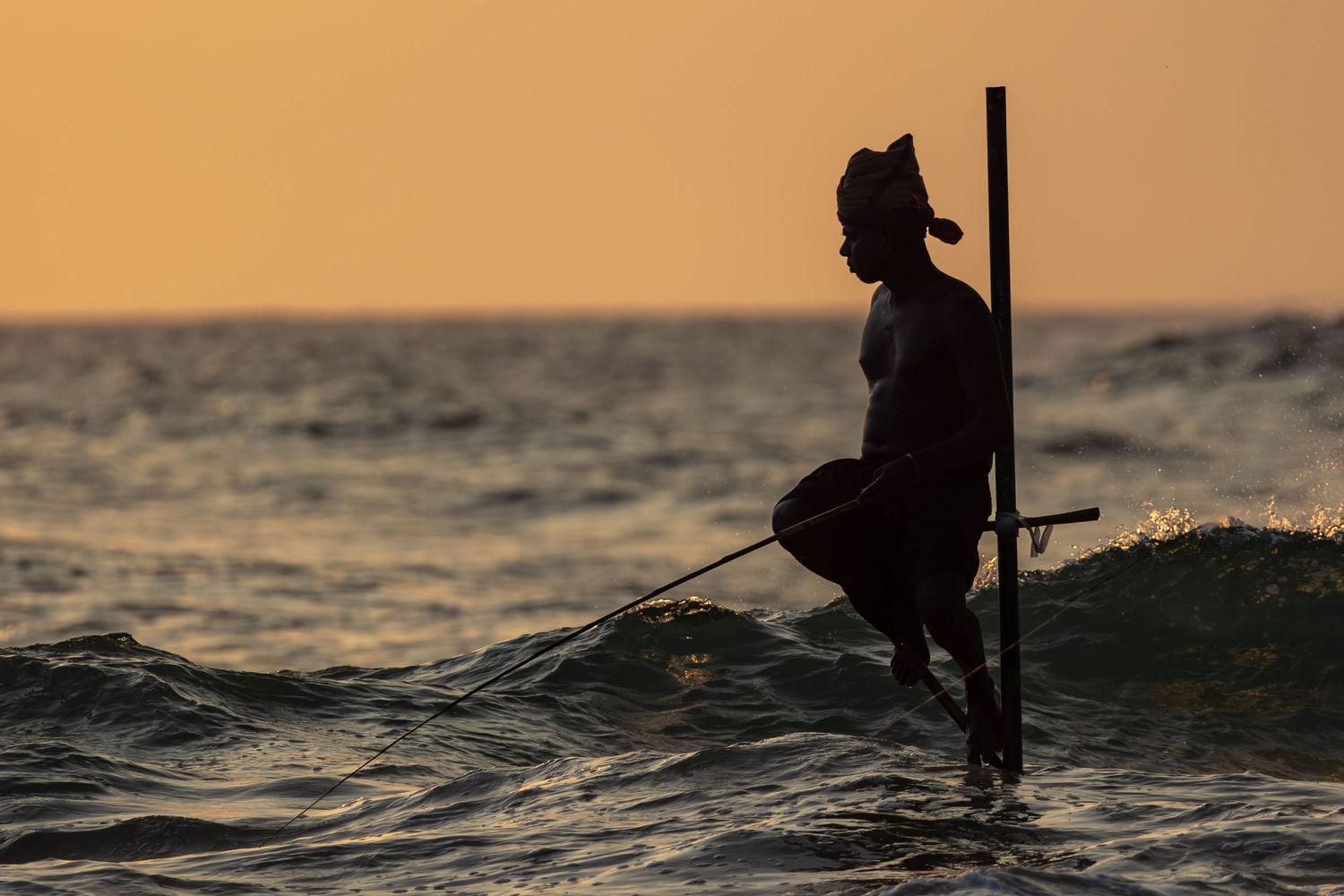 Silhouette of a traditional stilt fisher against an orange sunset sky.