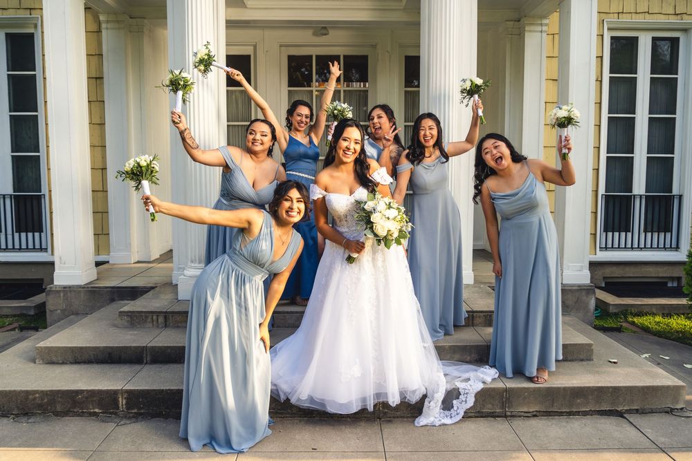 A joyful wedding party in light blue dresses poses enthusiastically on the steps of a the Lindley Scott House.